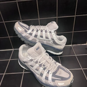 Nike P-6000 grå sneakers - Nike P-6000 sneakers i en snygg mix av ljusgrå och mörkgrå toner med vita detaljer. Ovandelen är i mesh och mocka med coola paneler och snörning. Sulan är robust och har bra grepp, perfekt för dig som gillar retrostil och sportig vibe.