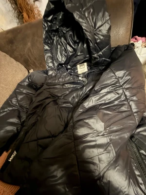 Svart pufferjacka från Warp - Svart pufferjacka från Warp med glansig yta och huva. Jackan har dragkedja framtill och två sidofickor med dragkedja. Quiltad design som ger en cool look och extra värme. Perfekt för kalla dagar och enkel att matcha med din stil.