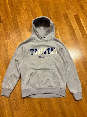Grå Trapstar hoodie med blå/vit text - Säljer en grå hoodie från Trapstar i storlek M med stor, fluffig blå och vit text på bröstet. Tröjan har huva med metallöljetter, magficka och ribbade muddar. Materialet är mjukt och fleecefodrat för extra komfort. Perfekt för chilliga dagar.