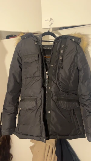 Svart parkajacka med päls från Hollies - Svart parkajacka från Hollies med snygg fuskpäls på huvan, flera praktiska fickor och detaljerade knappar. Jackan har dragkedja och tryckknappar framtill, samt patch med logga på ärmen. Perfekt för kalla dagar och har en klassisk, stilren look. Priset kan diskuteras 