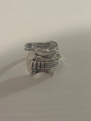 Silvrig ring med gitarrdesign - Unik silvrig ring formad som en elgitarr, perfekt för dig som älskar musik och vill sticka ut. Ringen har detaljerade strängar och knappar som ger den en cool och edgy vibe. Justerbar storlek så den passar de flesta fingrar. Pris kan diskuteras💕