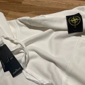 Vit hoodie från Stone Island - Vit hoodie från Stone Island med klassisk patch på ärmen och dragsko i huvan. Tröjan har känguruficka framtill och ribbade muddar vid ärmslut och midja. Perfekt för en clean och stilren look.