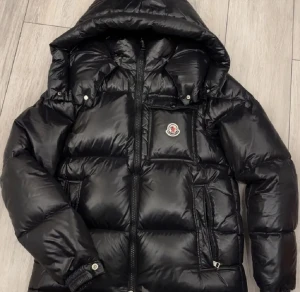 Svart Moncler pufferjacka med huva - Svart pufferjacka från Moncler med glansig finish, huva och ikonisk logga på bröstet. Jackan har flera fickor med dragkedja och quiltad design som ger en cool och sportig vibe. Perfekt för kalla dagar när du vill vara både varm och stilren.