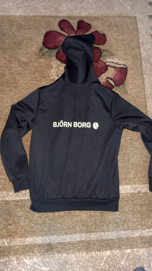 Svart hoodie med dragkedja Björn Borg - Svart hoodie från Björn Borg i storlek S, tillverkad av 95% polyester och 5% elastan. Tröjan har huva med snörning, dragkedja framtill och tryckt logga på bröstet. Perfekt för chill dagar eller när du vill ha en sportig look.