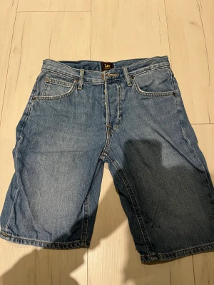 Blå jeansshorts från Lee - Klassiska blå jeansshorts från Lee med fem fickor, bälteshällor och Lee-lapp bak i midjan. Raka ben och normal passform, tillverkade i slitstark bomullsdenim. Perfekta för en avslappnad stil under varmare dagar.