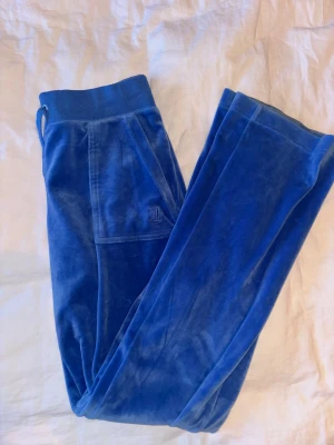 Blå mjukisbyxor från Juicy Couture - Snygga blå mjukisbyxor från Juicy Couture i mjuk sammet med broderad JC-logga på sidan. Byxorna har resår och snörning i midjan, samt bakfickor med knappar som har logodetalj. Perfekta för chill och sköna dagar. Byxorna är pyttelite missfärgade ner till, se sista bilden men utöver det så är det inga skavanker! Skriv gärna om du har någon fråga💞