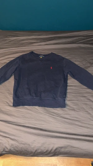 Mörkblå sweatshirt från Polo Ralph Lauren - Snygg mörkblå sweatshirt från Polo Ralph Lauren med klassisk rund hals och långärmad passform. Tröjan har det ikoniska röda Polo-logot på bröstet och är gjord i mjuk bomull. Perfekt för chill dagar och enkel att matcha med jeans eller joggers. Storlek M (10–12 år). I fint skick, inga hål eller fläckar. Pris kan diskuteras.