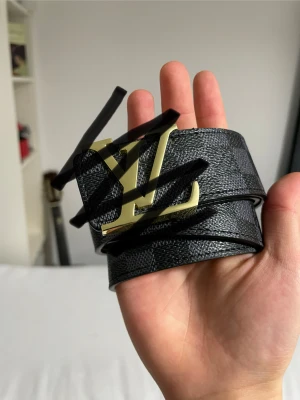 svart skinnbälte med guldspänne - Exklusivt bälte i svart skinn med diskret rutigt mönster och ikoniskt spänne i guld. Insidan är ljusbrun och har präglad logga. Perfekt accessoar för att lyfta din outfit med lyxig vibe.