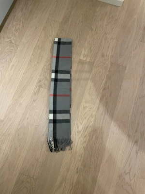 Bureberry halsduk - Snygg burberry i grått med klassiskt rutigt mönster i svart, vitt och rött. Mjukt material och fransar i ändarna ger en chill vibe. Perfekt att slänga över axlarna när det är kallt ute.