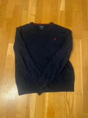 Mörkblå tröja från Polo Ralph Lauren - Snygg mörkblå tröja från Polo Ralph Lauren med klassisk röd logga broderad på bröstet. Rund hals med röd detalj på insidan av kragen. Tillverkad i mjuk pima bomull och har långa ärmar samt ribbade muddar vid ärmslut och nederkant.