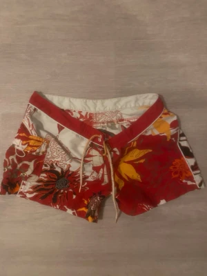 Färgglada blommiga shorts med snörning - Säljer ett par coola shorts med blommigt mönster i rött, gult, orange och vitt. De har låg midja, snörning framtill och praktisk ficka på sidan. Perfekta för sommaren och strandhäng. Materialet känns som syntet, snabbtorkande och lätt.