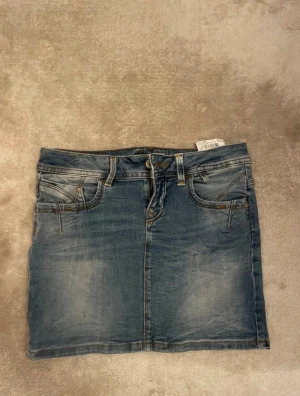 Blå jeanskjol från LTB - Snygg blå jeanskjol från LTB med klassisk femficksdesign, bälteshällor och knappstängning framtill. Kjolen har en rak passform och är i mjukt denimtyg med slitna detaljer för en cool look. Perfekt till sneakers eller boots.