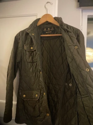 Olivgrön quiltad Barbour jacka - Quiltad olivgrön jacka från Barbour med snygga guldfärgade knappar och flera fickor framtill. Klassisk krage och figurnära passform. Jackan har lång ärm och är tillverkad i polyester, perfekt för dig som gillar stilren och tidlös design.