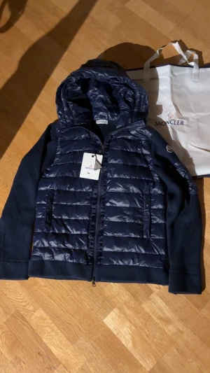 Mörkblå/Marinblå Moncler Cardigan NFC Scan - Snygg marinblå pufferjacka från Moncler med stickade ärmar och huva. Jackan har quiltad front i glansig polyester, dragkedja och Moncler-logga på ärmen. Storlek L men passar helst dem med M. Helt ny, alltså aldrig använd. Skriv om ni har frågor.  NFC SCAN!