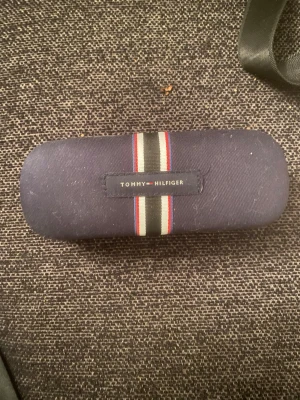Blå glasögonfodral Tommy Hilfiger - Snyggt glasögonfodral från Tommy Hilfiger i mörkblått tyg med klassiska Hilfiger-ränder i rött, vitt och svart. Fodralet är avlångt och har en mjuk, tygklädd utsida med logotypen framtill. Perfekt för att hålla dina glasögon safe och stilrena.
