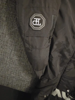 Svart pufferjacka från TPDY - Säljer en svart pufferjacka från TPDY med huva och coola vita tryckdetaljer på bröstet och ärmen. Jackan har flera fickor med dragkedja och knappar, samt ett snyggt märke på ärmen. Perfekt för kalla dagar och streetwear-stil.