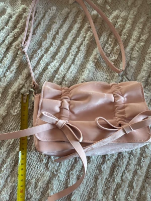 Rosa axelväska med rosetter - Söt ljusrosa axelväska med justerbar rem och två dekorativa rosetter framtill. Väskan har ett quiltat lock med magnetknäppning och är tillverkad i mjuk mocka. Perfekt storlek för det viktigaste och ger en gullig touch till din outfit.