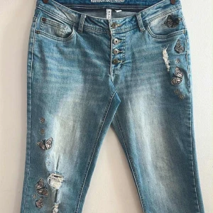 Blå boyfriend-jeans med fjärilar - Snygga ljusblå boyfriend-jeans från Rainbow med broderade fjärilar och blommor på benen. Jeansen har distressed detaljer, knappar i midjan och klassisk femficksdesign. Perfekt för dig som gillar en chill och kreativ stil.