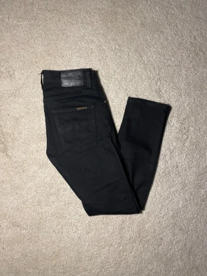 Svarta Nudie Jeans 28/30 grim trim - Svarta jeans från Nudie Jeans med klassiska fem fickor och svarta sömmar. Tillverkade av 99% ekologisk bomull och 1% elastan för extra stretch. Snyggt minimalistiskt utseende med svart läderpatch bak i midjan. Modellen är grim trim 