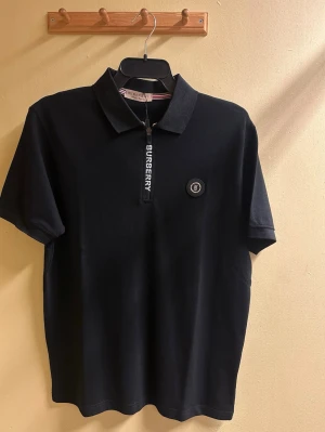 Svart polotopp från Burberry - Svart polotopp från Burberry med korta ärmar och klassisk krage. Toppen har en snygg dragkedja framtill med Burberry-tryck och ett rundat emblem med B-logga på bröstet. Materialet är mjukt och känns lyxigt, perfekt för dig som gillar stilrena designer.