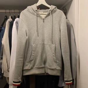 Grå hoodie från Moncler - Säljer en stilren grå hoodie från Moncler med dragkedja och huva. Tröjan har ribbade muddar och detaljer i rött, vitt och blått på ärmslut och huva. Knappt använd! Storlek S med väldigt bra skick. Passar till 170-180