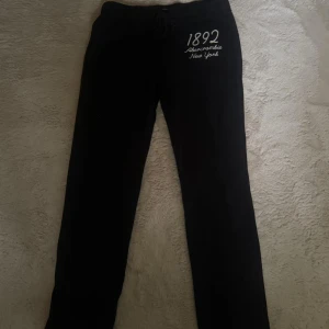 Svarta mjukisbyxor Abercrombie M - Svarta mjukisbyxor från Abercrombie & Fitch med broderad vit text '1892 Abercrombie New York' på ena benet. Byxorna har snörning i midjan och är tillverkade i mjukt material som passar perfekt för chill dagar.