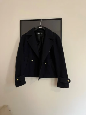 Marinblå skepparkavaj från Zara - Snygg marinblå skepparkavaj från Zara med breda slag och guldknappar. Jackan har två fickor fram och dekorativa knappar vid ärmsluten. Perfekt för dig som gillar klassisk stil med en modern twist.