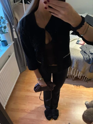 Svart glittrig kofta - Säljer en svart glittrig kofta med trekvartsärm och dekorativa dragkedjor vid fickorna. Koftan har en öppen front och är perfekt för dig som vill ha en chic och trendig look. Materialet är mjukt och har en lätt glans som fångar ljuset snyggt.