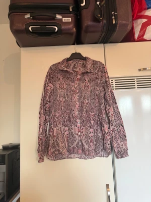 Mönstrad rosa skjorta från Odd Molly - Säljer en unik långärmad skjorta från Odd Molly i ljusrosa och vinröda toner med ett detaljerat grafiskt mönster. Skjortan har knappar framtill, klassisk krage och är tillverkad i ett tunt, luftigt material som känns riktigt skönt på. Perfekt för dig som gillar statement-plagg! Från märket Odd Molly. Storlek 2 som enligt deras storleksguide motsvarar en M. Mycket fint skick, jag hittar inga synliga anmärkningar eller defekter.  