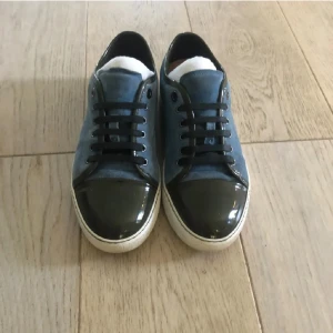 Blå mockasneakers från Lanvin - Snygga sneakers från Lanvin i blå mocka med svarta lackdetaljer på tån och svarta skosnören. Skorna har vit sula och klassisk låg profil. Kommer med originalkartong och dustbag. Perfekta för dig som gillar exklusiva och stilrena sneakers.