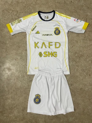 Al Nassr Ronaldo matchställ Adidas - Säljer ett Al Nassr matchställ med Ronaldo 7-tryck. Setet består av vit tröja och shorts med gula detaljer och klubbmärke. Tröjan har svarta och gula inslag, tryckta sponsorer och Aeroready-material för extra komfort. Perfekt för fotbollsfans!
