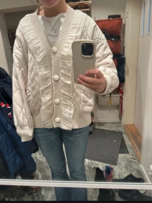 Beige cardigan jacka med stora knappar - Superfin beige cardigan jacka med quiltad struktur och stora, matchande knappar framtill. Jackan har v-ringning, ribbade muddar och ett coolt rutmönster i materialet. Perfekt för dig som gillar en mysig och trendig look.