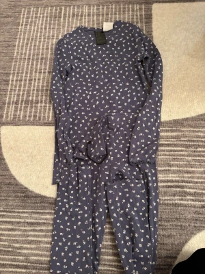 Blommig blå byxdress från H&M - Säljer en blå byxdress från H&M med vita blommor och ribbad struktur. Byxdressen har långa ärmar, rund hals och ett matchande midjeband. Perfekt för dig som gillar avslappnad och söt stil.