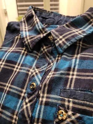 Blå rutig skjorta med guldknappar - Snygg blå och mörkblå rutig skjorta med guldiga knappar och bröstficka. Skjortan har långa ärmar och förstärkta axelpartier i mörkare tyg. Perfekt för dig som gillar en avslappnad men ändå stilren look.