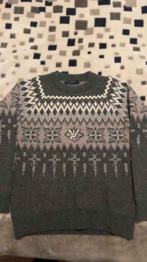 Grå Louis Vuitton stickad tröja - Säljer en grå stickad tröja från Louis Vuitton med snyggt vintermönster i vitt och beige. Tröjan har rund hals, långa ärmar och LV-logga mitt på bröstet. Perfekt för kalla dagar när du vill vara både varm och stilren. Prisets kan diskuteras.