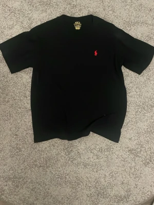 Svart t-shirt från Polo Ralph Lauren - Svart t-shirt från Polo Ralph Lauren med klassisk röd broderad logga på bröstet. T-shirten har rund halsringning och korta ärmar. Tillverkad i mjuk bomull för en skön känsla och enkel stil.