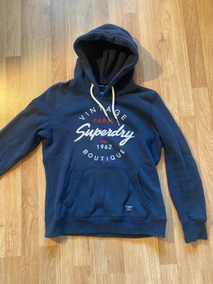 Superdry hoodie marinblå - Supermysig marinblå hoodie från Superdry med stort tryck framtill. Har några fläckar framtill som syns, har använt en del men inga andra defekter ( se bilder för egen bedömning). Jätte mysig och bekväm. Är i stolek EU40 men skulle mer säga att den är EU38. Hör av er vid frågor!💕