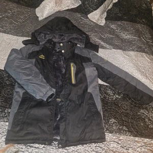 Svart och grå vindjacka Outdoor Sport - Svart och grå vindjacka från Outdoor Sport med huva och mjukt fleecefoder. Jackan har en gul detalj på bröstet, dragkedja och flera fickor. Perfekt för kalla dagar och har en sportig look med broderad logga på bröstet. Helt ny oanvänd, endast testad. Finns två! 1 för 300 eller båda för 500, meddela isåfall innan köp av 2.