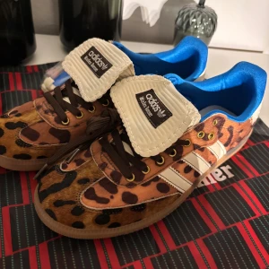 Adidas Samba leopardmönstrade sneakers - Unika Adidas Samba sneakers med leopardmönster i bruna och svarta toner, vita detaljer och blå insida. Skorna har klassiska vita ränder, bruna snören och gummisula. Materialet är skinn med pälsstruktur och extra snören medföljer. Perfekt för dig som vill sticka ut.