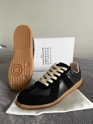 Maison Margiela skor  - Maison Margiela sneakers i svart skinn och mocka med beige snörning och gummisula i naturfärg. Klassisk rund tå och platt sula, med ikonisk etikett på tungan. Snygg och stilren design som passar till streetwear och modeintresserade.