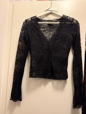 Svart spetsblus från Gina Tricot XS - Supersnygg svart blus i spets från Gina Tricot. Transparent med blommigt spetsmönster, v-ringning och knappar framtill. Långärmad och figurnära passform, perfekt att styla lager på lager eller som statement-piece.