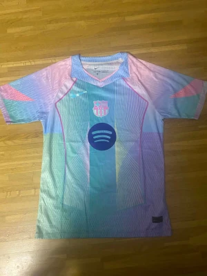 Pastellfärgad FC Barcelona Nike tröja - Säljer en unik FC Barcelona tröja från Nike i pastellfärger med inslag av blått, rosa, lila och grönt. Tröjan har klubbmärke och sponsortryck framtill samt ett futuristiskt mönster. Perfekt för dig som vill sticka ut på läktaren eller i vardagen.