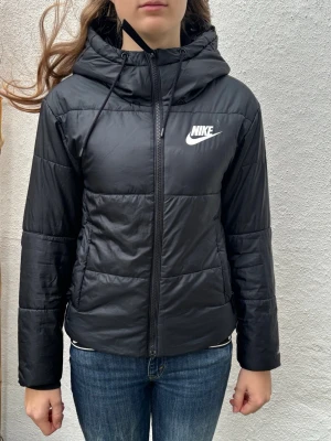 Svart pufferjacka Nike Therma-Fit XS - Svart pufferjacka från Nike i modellen Therma-Fit. Jackan har huva med snörning, dragkedja framtill och Nike-logga på bröstet. Lätt vadderad och perfekt för kyliga dagar. Snygg och sportig look som passar till jeans eller träningskläder.