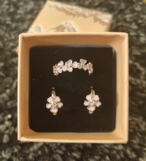 Blommiga smyckeset  - Ett elegant set från Wostu med örhängen och ring i sterling silver (stämplade 925). • Ringstorlek: 7 (54) • Örhängen: diameter 1,3 cm, omkrets 4 cm (barnstorlek) OBS! Oxidation kan uppstå med tiden