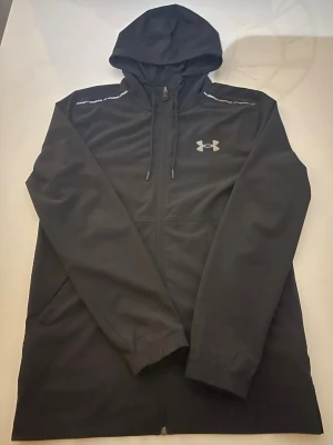 Under Armour Jacka Herr - Svart Under Armour jacka , Herr jacka. Jackan är ganska lång för och vara s och har en sinched midja. Axlarna har text på sig med under armor och är reflekterande. Jackan är ganska ny och tvättas och skickas.