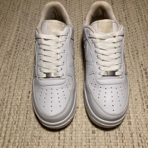 Nike Air Force 1 vita sneakers - Fräscha Nike Air Force 1 sneakers i helvitt skinn med klassisk lag siluett. Snygga perforeringar på tån, vit sula och snörning med metall-detalj. Ikonisk Swoosh på sidan och mjuk vadderad insida för extra komfort. Perfekt för dig som gillar cleana och stilrena sneakers.