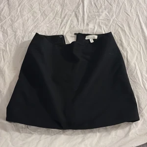 Svart kortkjol från H&M, strl 36 - Snygg svart kortkjol från H&M i klassisk A-linjeform. Kjolen har en diskret dragkedja i sidan och är tillverkad i ett slätt material som ger en stilren look. Perfekt att matcha med en cool topp eller hoodie. Passar dig som gillar enkel och clean stil.