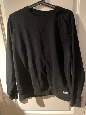 Svart sweatshirt från 157, stl S - En stilren svart sweatshirt från 157 Denim Makers med rund hals och ribbade muddar. Tröjan är i mjuk bomull och har en liten vit etikett nertill. Perfekt för dig som gillar enkel och clean stil.