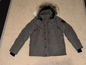 Grå dunjacka Canada Goose med pälskrage - Snygg grå dunjacka från Canada Goose med svart logotyp på ärmen och fluffig pälskant på huvan. Jackan har dragkedja och knappar framtill, långa ärmar med muddar och flera praktiska fickor. Perfekt för kalla vinterdagar.