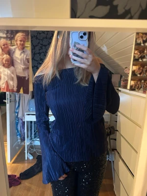Mörkblå plisserad blus med trumpetärm - Säljer en mörkblå blus med plisserat tyg och breda trumpetärmar. Blusen har en rak, lite offshoulder halsringning och är tillverkad i ett glansigt syntetmaterial som ger en cool vibe. Perfekt för dig som gillar unika detaljer och vill sticka ut.